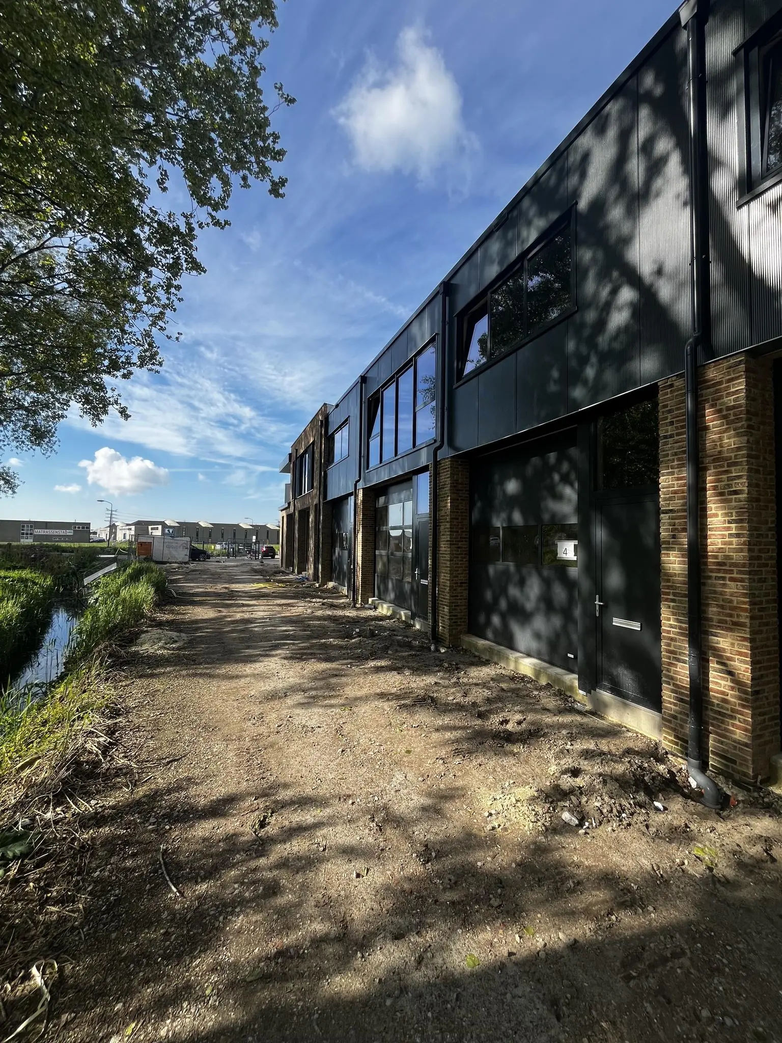 Nieuwbouwpand aan Broekdijk West met industriële gevel en onverharde oprit langs een sloot.